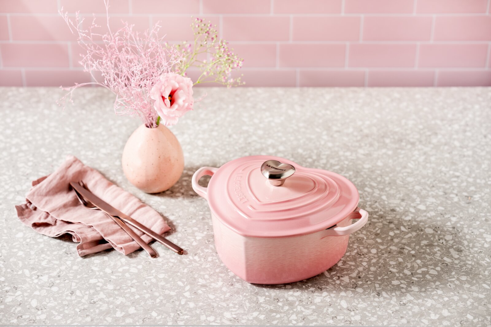 Rosa, herzförmiger Le Creuset Gusseisentopf mit Deckel und Edelstahlknauf auf heller Terrazzo-Arbeitsplatte, links daneben rosafarbenes Besteck auf Stoffserviette und eine kleine Vase mit rosa Blumen; Perspektive: leicht erhöhte Frontansicht (3/4).