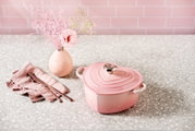 Rosa, herzförmiger Le Creuset Gusseisentopf mit Deckel und Edelstahlknauf auf heller Terrazzo-Arbeitsplatte, links daneben rosafarbenes Besteck auf Stoffserviette und eine kleine Vase mit rosa Blumen; Perspektive: leicht erhöhte Frontansicht (3/4).