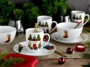 Weihnachtliches Porzellan-Geschirrset mit Tasse auf Untertasse und festlichen Strumpf- und Ornamentmotiven, dekoriert mit goldfarbenem Löffel, Kerze und roten Kugeln auf Holztisch vor Tannenzweigen, Perspektive: leicht seitliche Frontansicht auf Tischhöhe