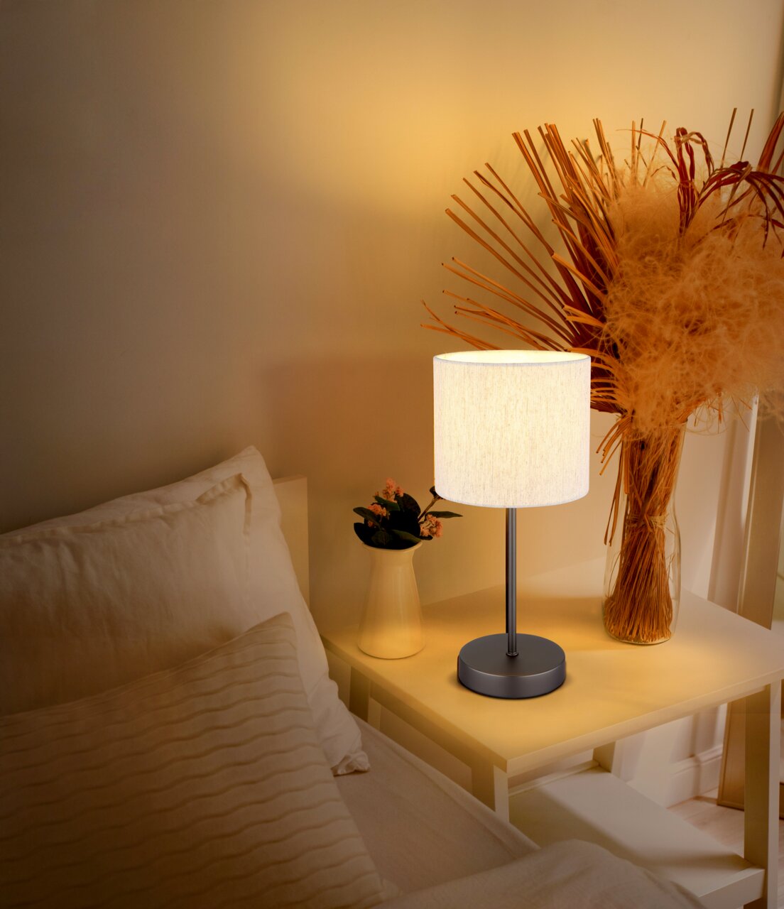 Tischleuchte mit weißem Lampenschirm auf einem Nachttisch, umgeben von Dekorationen, aus seitlicher Perspektive fotografiert.