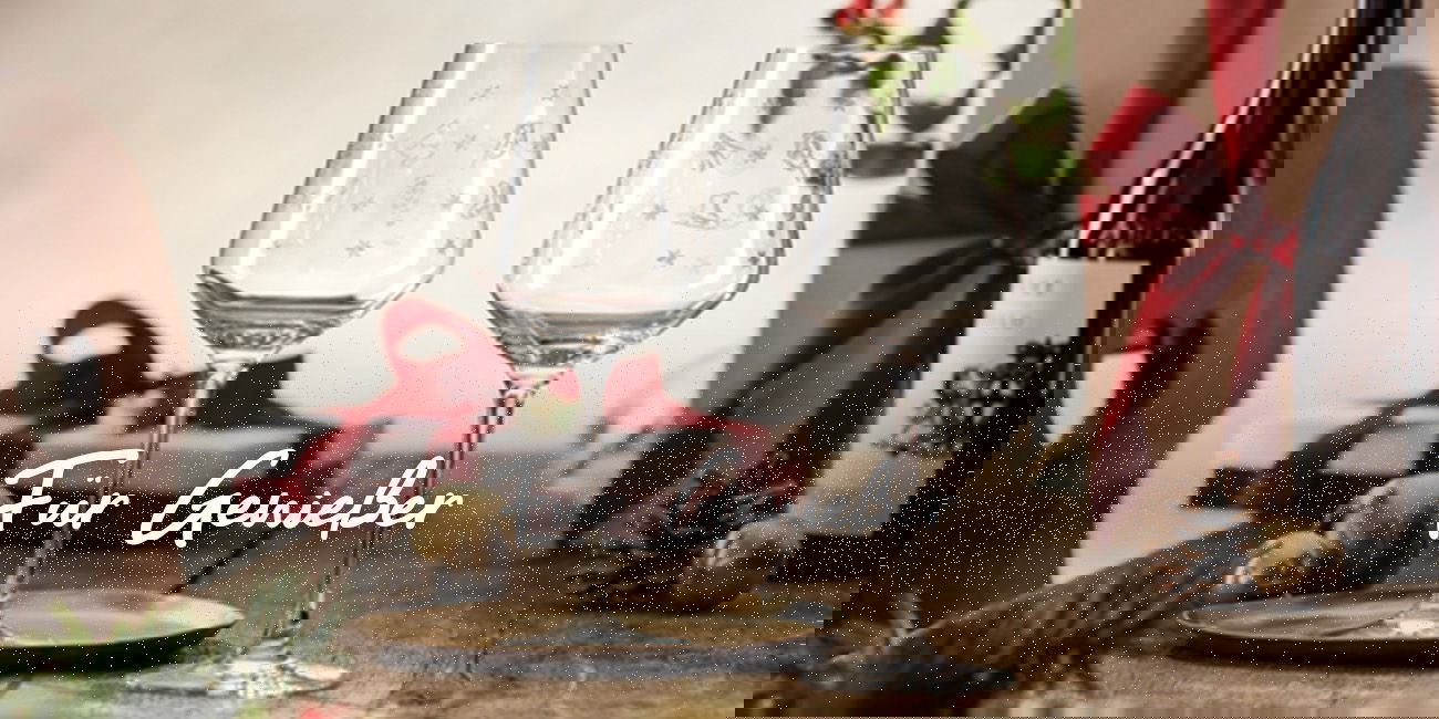 Zwei festliche Weingläser auf Holztisch mit Geschenk und Weinflasche, darüber Schriftzug „Für Genießer“.