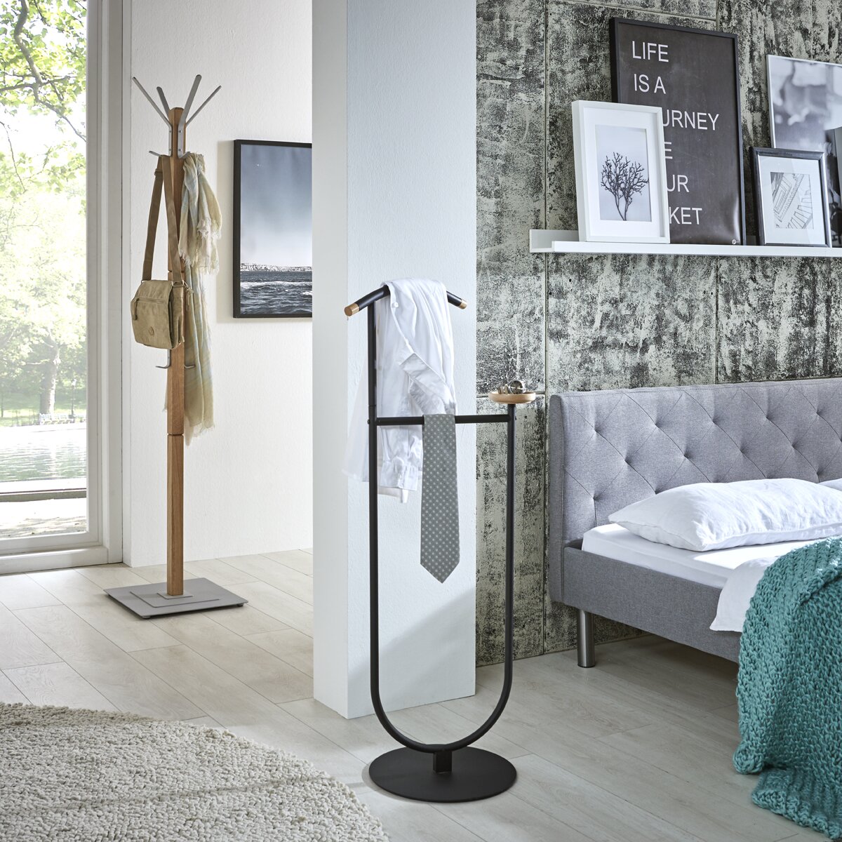 Schwarzer Kleiderständer/Butler mit rundem Standfuß, Holzdetails und Ablage, mit Hemd und Krawatte im Schlafzimmer neben grauem Bett, Perspektive: leicht seitliche Frontansicht