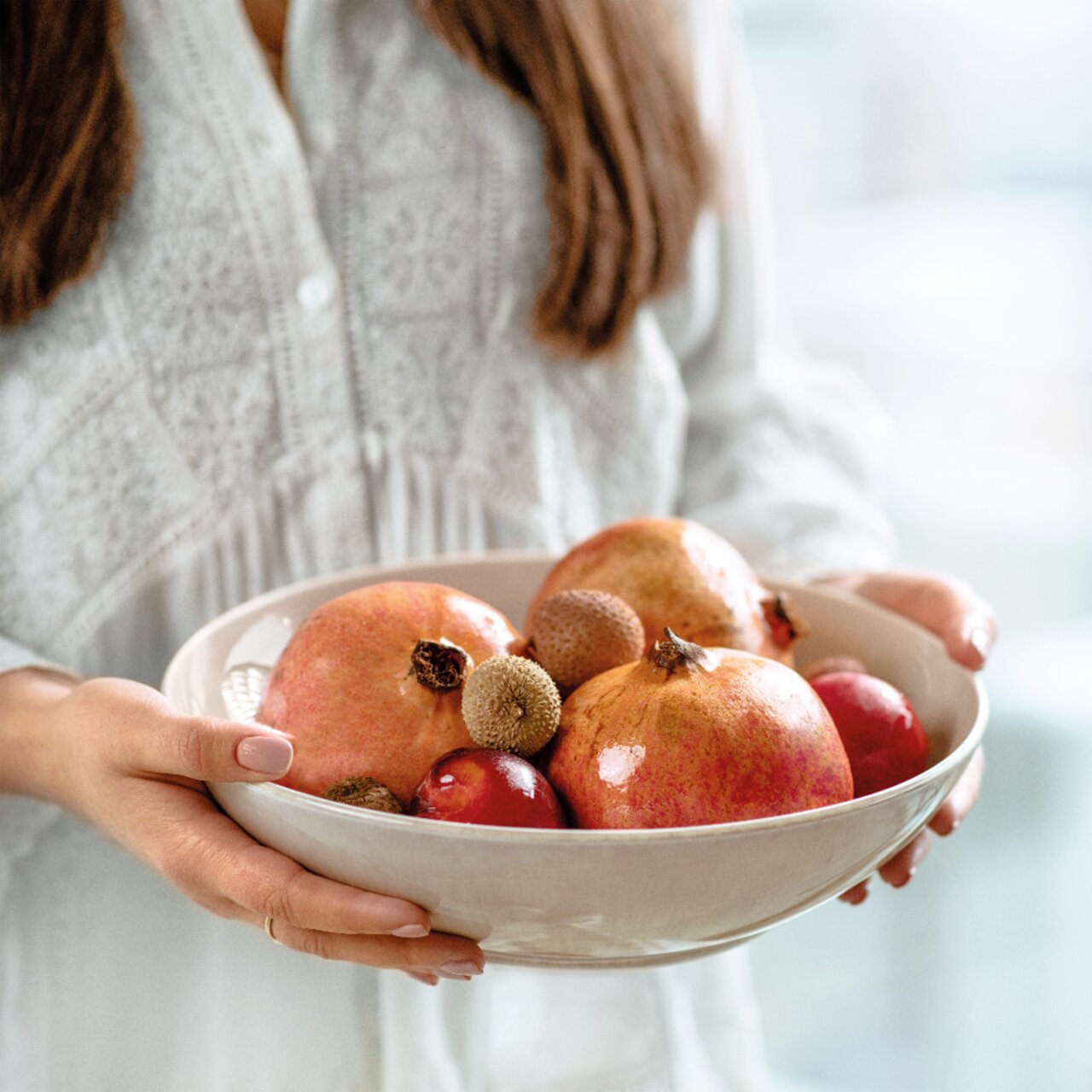 Eine Frau hält eine cremefarbene Schale mit Granatäpfeln und anderen Früchten, seitliche Perspektive.
