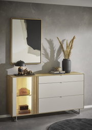 Modernes Sideboard aus Holz mit hellen Fronten, drei Schubladen und beleuchtetem offenen Fach, dekoriert mit Lampe, Vase und Büchern, frontal leicht schräg von links fotografiert.