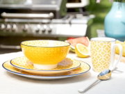 Gelb-weißes Geschirrset mit geometrischem Muster: Schüssel auf zwei Tellern, daneben passende Tasse und Löffel auf einem Tisch im Freien, fotografiert aus leicht schräger Vorderansicht auf Augenhöhe.