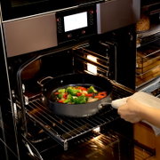 Hand schiebt eine große schwarze Bratpfanne mit Gemüse (Brokkoli, Paprika, Zwiebeln) in einen geöffneten modernen Backofen mit Display und Innenbeleuchtung; Perspektive: seitliche Nahaufnahme von vorn rechts.