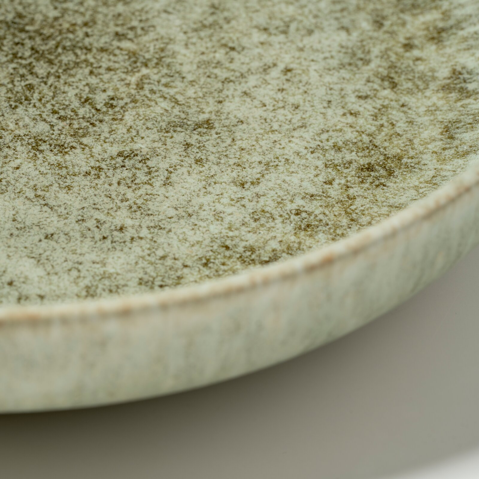 Detailaufnahme aus schräger Seitenperspektive: gesprenkelter, hellgrüner Keramikteller/Schale mit rauer, glasiert wirkender Oberfläche und sichtbarem Rand