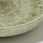 Detailaufnahme aus schräger Seitenperspektive: gesprenkelter, hellgrüner Keramikteller/Schale mit rauer, glasiert wirkender Oberfläche und sichtbarem Rand