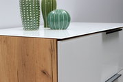 Nahaufnahme einer Schubkastenkommode mit weißer Hochglanzoberfläche und Holzdekor, dekoriert mit grünen Vasen. Perspektive von der Seite.