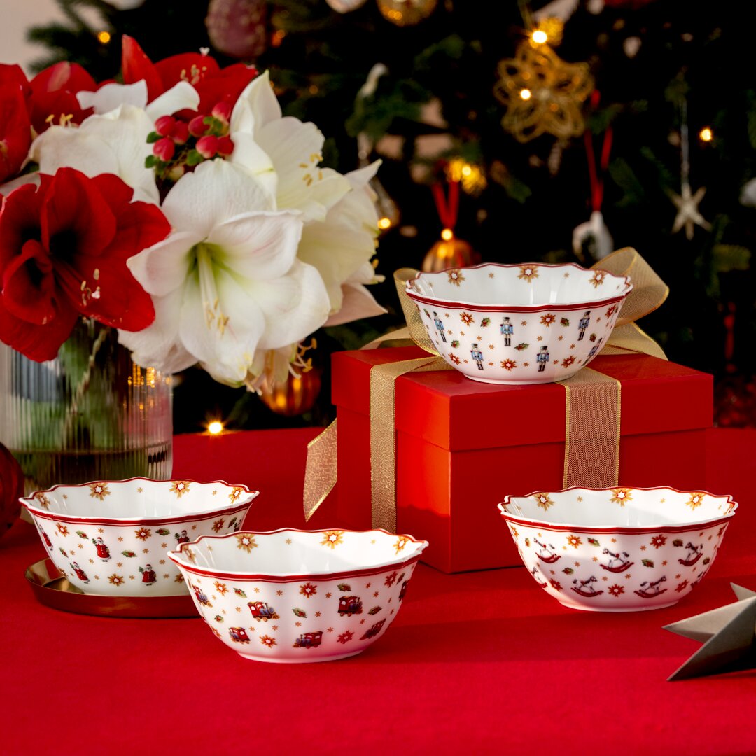 Villeroy & Boch Jubiläums Schalen-Set 4tlg Vier festliche Schalen mit weihnachtlichen Motiven, präsentiert auf einem Tisch mit rotem Tuch, im Hintergrund ein geschmückter Weihnachtsbaum und ein Blumenarrangement. Perspektive: seitlich von vorne.