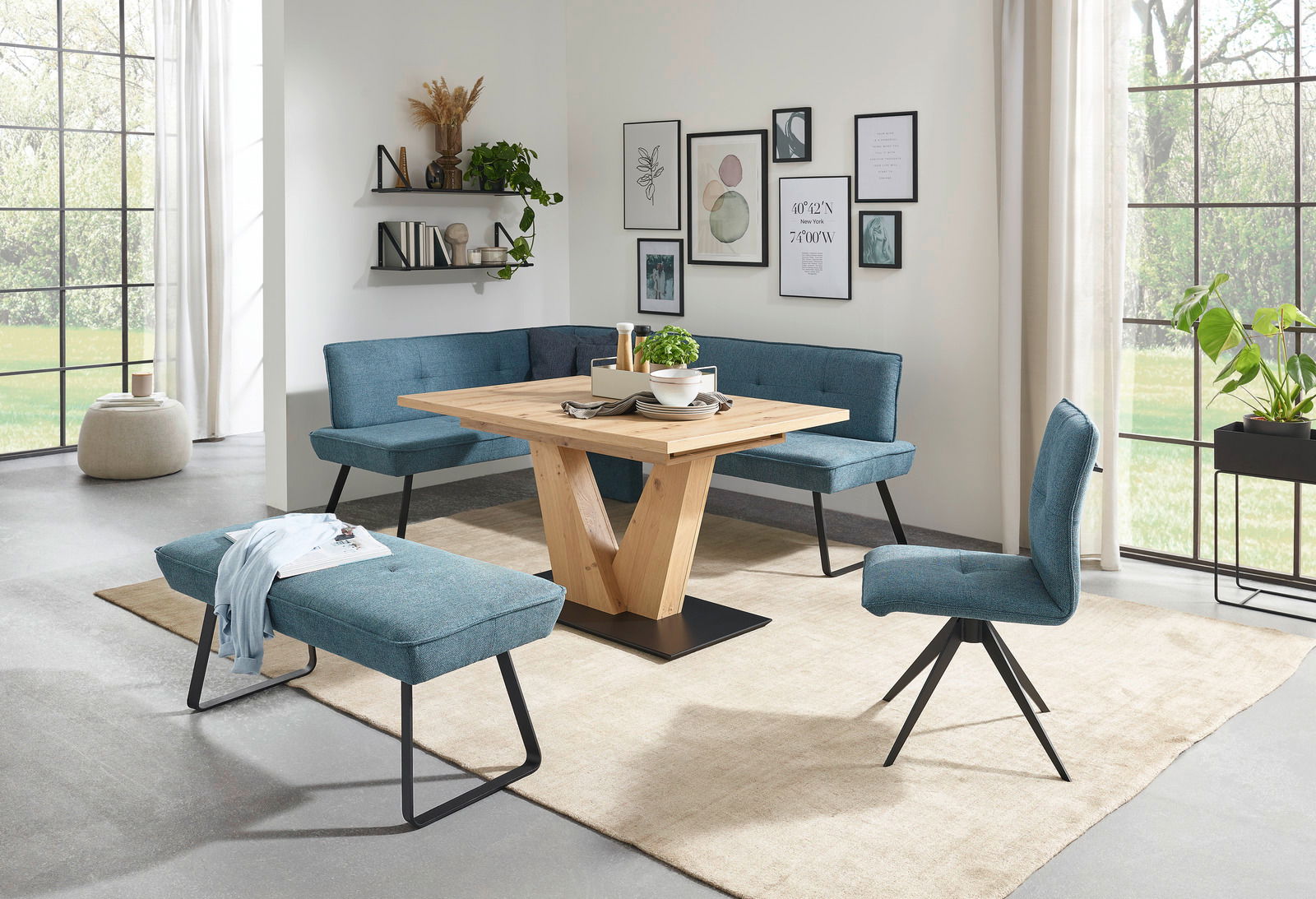 Moderne Esszimmer-Einrichtung mit blauer Eckbank, passendem Stuhl und Hockerbank, sowie einem Holztisch mit V-Gestell, aus der Perspektive des Raumes betrachtet.