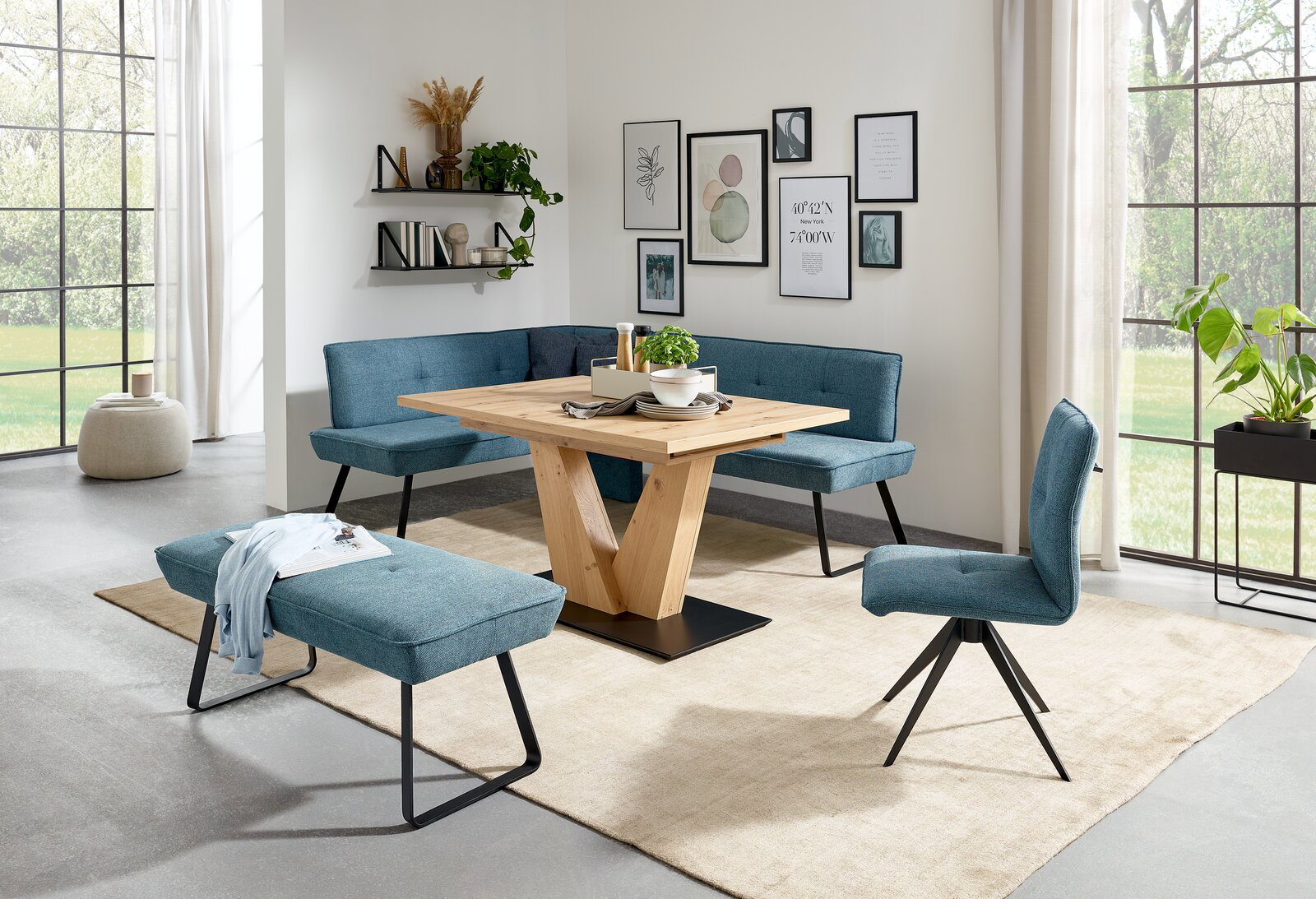 Moderne Esszimmer-Einrichtung mit blauer Eckbank, passendem Stuhl und Hockerbank, sowie einem Holztisch mit V-Gestell, aus der Perspektive des Raumes betrachtet.