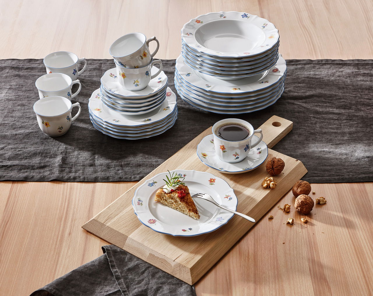 Kombiservice Sonate 30-tlg. mit floralem Muster, bestehend aus Tellern, Tassen und Untertassen, auf einem Tisch mit Holzbrett und Nüssen, aus der Vogelperspektive fotografiert.