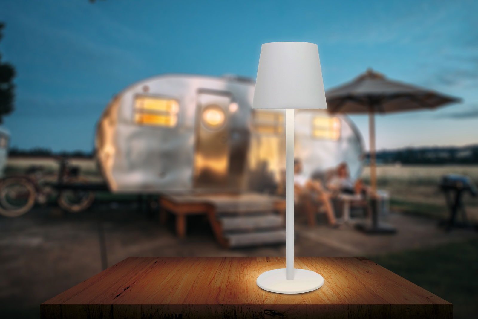Weiße Akku-Tischleuchte auf einem Holztisch, im Hintergrund ein unscharfer Wohnwagen und eine Terrasse bei Dämmerung, seitliche Perspektive.