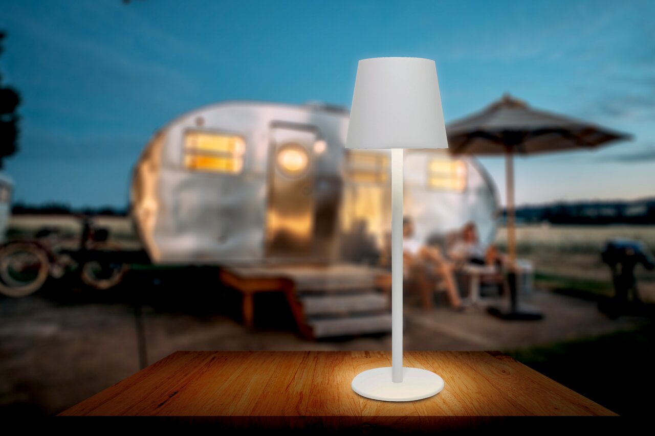 Weiße Akku-Tischleuchte auf einem Holztisch, im Hintergrund ein unscharfer Wohnwagen und eine Terrasse bei Dämmerung, seitliche Perspektive.