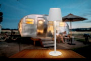 Weiße Akku-Tischleuchte auf einem Holztisch, im Hintergrund ein unscharfer Wohnwagen und eine Terrasse bei Dämmerung, seitliche Perspektive.