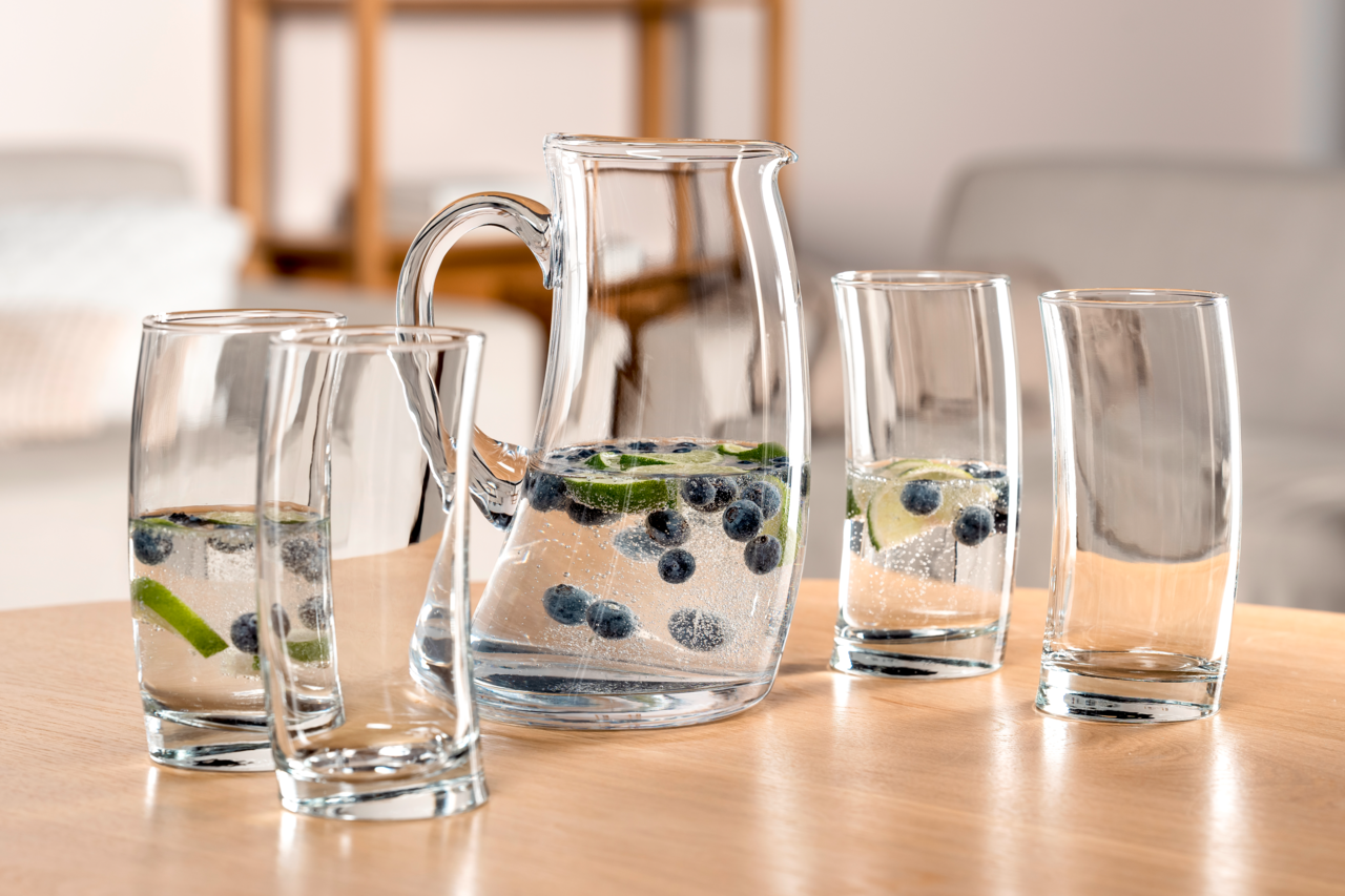 Set aus einem Krug und vier Gläsern, gefüllt mit Wasser, Limettenscheiben und Blaubeeren, auf einem Tisch in einer gemütlichen Wohnumgebung, seitliche Perspektive.