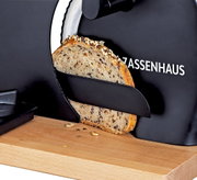 Nahaufnahme aus seitlicher Perspektive: Schwarze Brotschneidemaschine von Zassenhaus schneidet eine Scheibe Körnerbrot auf einem Holzbrett.