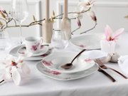 Elegant gedeckter Tisch mit weißem Porzellan-Geschirrset mit rosa Magnolienblüten-Dekor, Tasse, Teller und Schale sowie Glas und roségoldfarbenem Besteck auf weißer Tischdecke, aufgenommen aus leicht erhöhter schräger Vorderansicht.