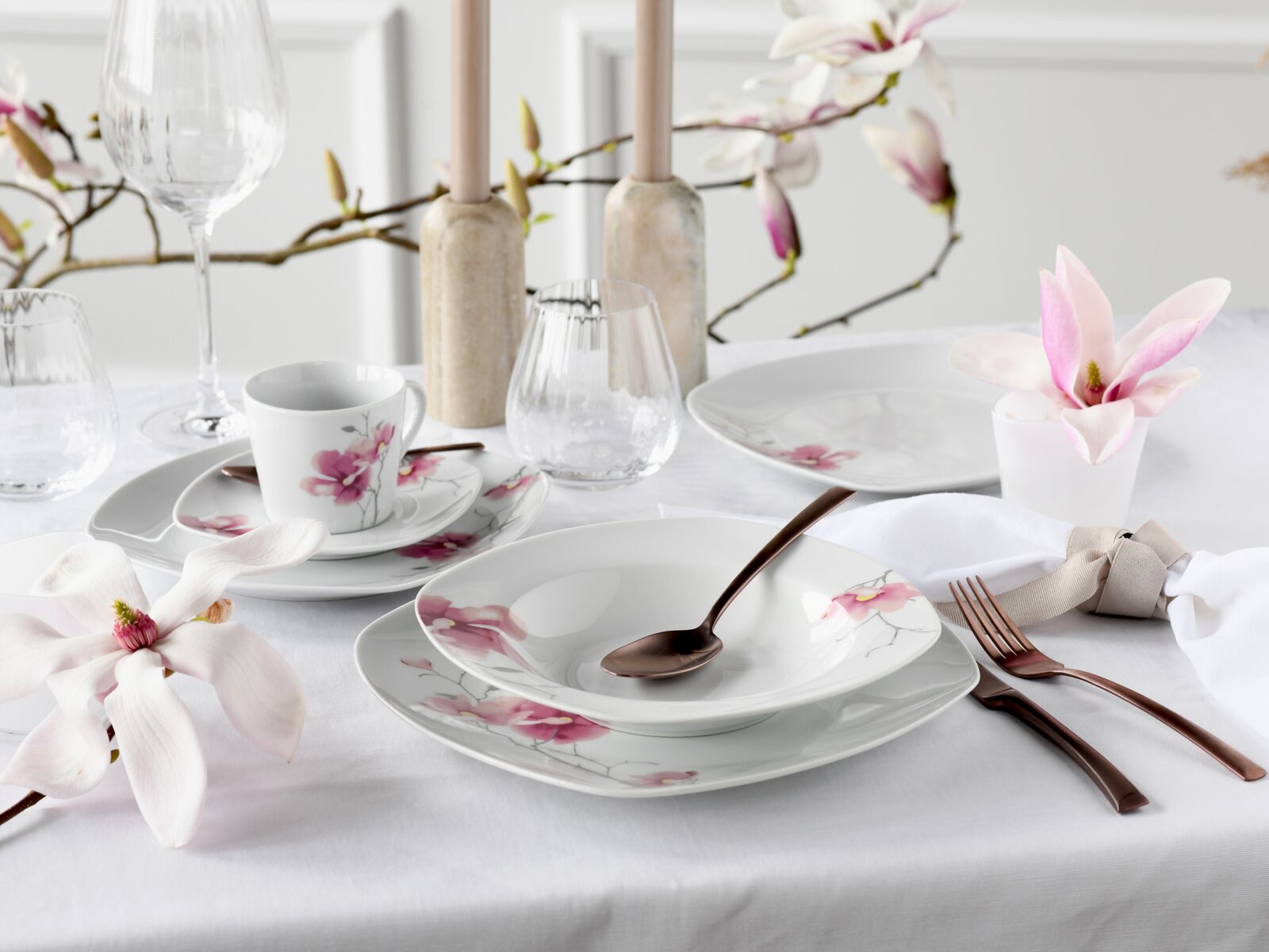 Elegant gedeckter Tisch mit weißem Porzellan-Geschirrset mit rosa Magnolienblüten-Dekor, Tasse, Teller und Schale sowie Glas und roségoldfarbenem Besteck auf weißer Tischdecke, aufgenommen aus leicht erhöhter schräger Vorderansicht.