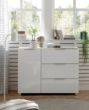 Weiße Kommode mit Hochglanzoberfläche, zwei Türen und zwei Schubladen, frontal fotografiert, dekoriert mit Pflanzen und Büchern, vor einem Fenster mit Blick ins Grüne.