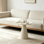 Minimalistischer runder Beistelltisch in Cremeweiß mit konischem Fuß und Tablettkante, im Wohnzimmer vor einem hellen Sofa, fotografiert in frontaler leicht seitlicher Perspektive auf Tischhöhe