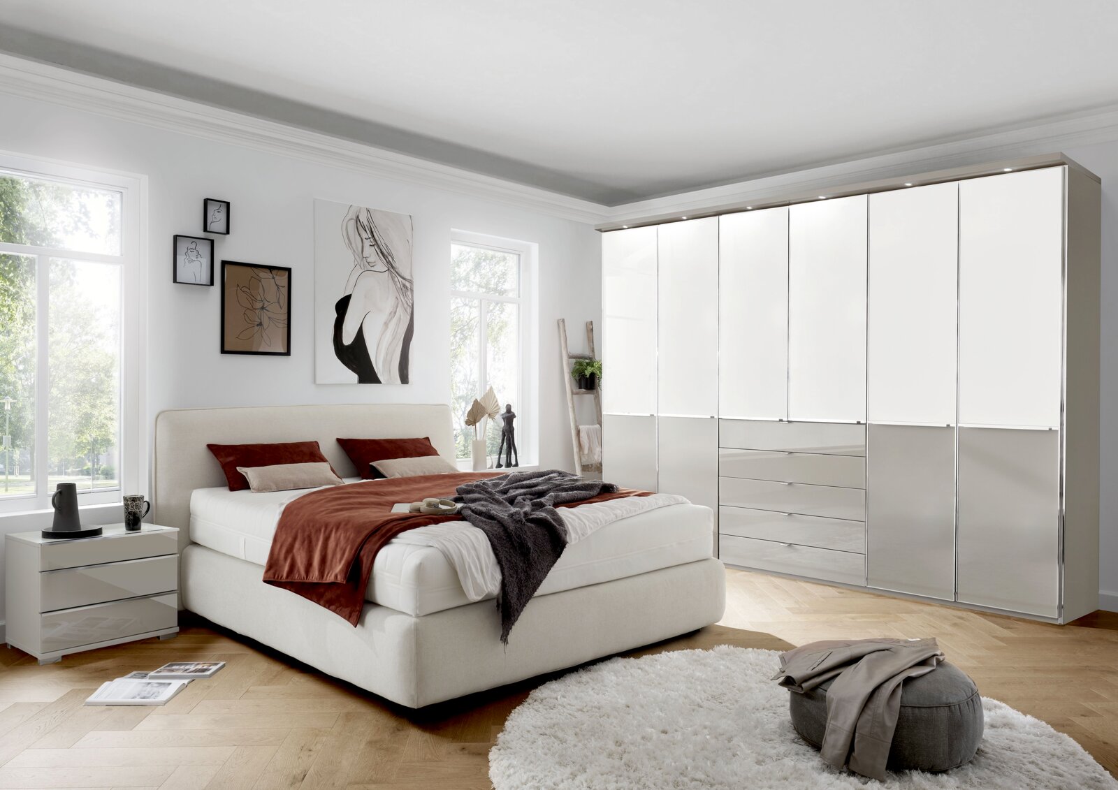 Modernes Schlafzimmer mit gepolstertem Boxspringbett in Creme, Nachttisch und großem weißen Kleiderschrank mit Hochglanzfronten und Schubladen, aufgenommen in schräger Frontansicht aus Augenhöhe.