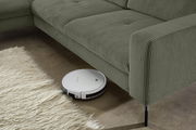 Ein Saugroboter reinigt den Boden unter einem grünen Longchair aus geripptem Stoff, Perspektive von der Seite.