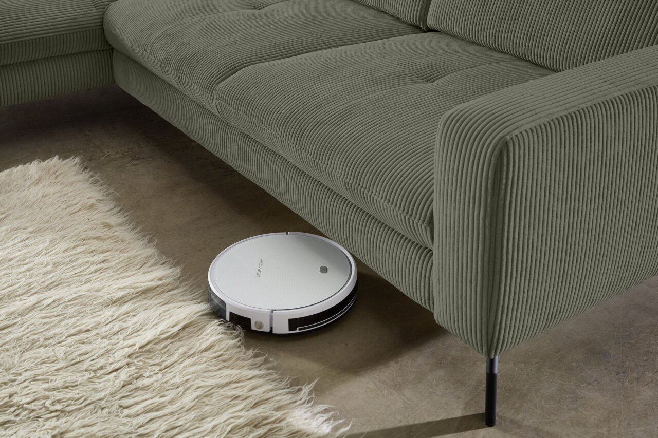 Een robotstofzuiger maakt de vloer schoon onder een groene ligstoel van geribbelde stof, gezien vanaf de zijkant.