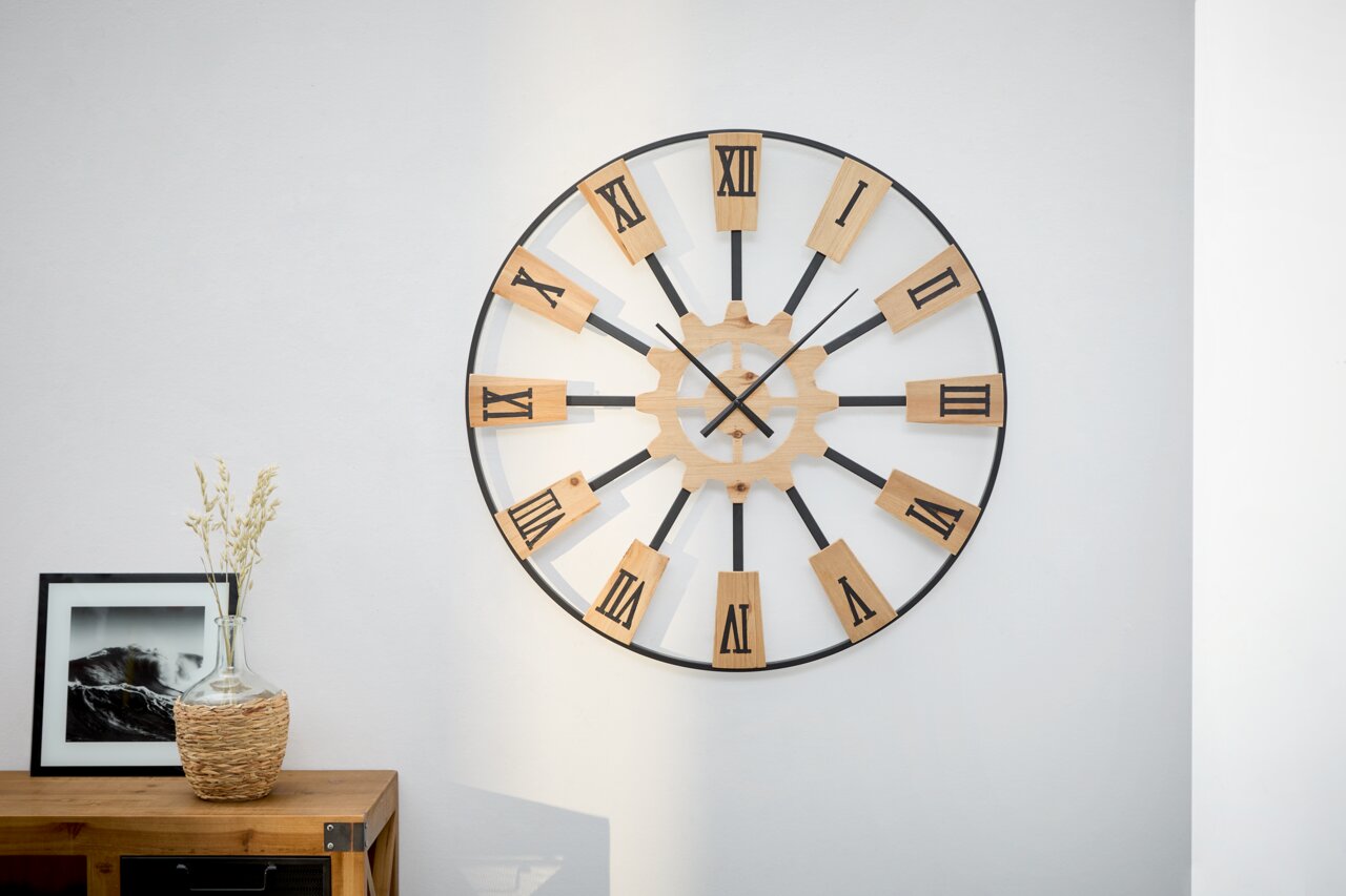 Große Wanduhr mit römischen Ziffern aus Holz und Metall, frontal an einer weißen Wand montiert.