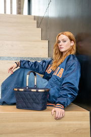 Junge Frau sitzt auf einer Treppe und lehnt sich an die Wand, neben ihr steht eine kleine, gesteppte schwarze Tasche mit hellen Griffen. Perspektive von vorne.