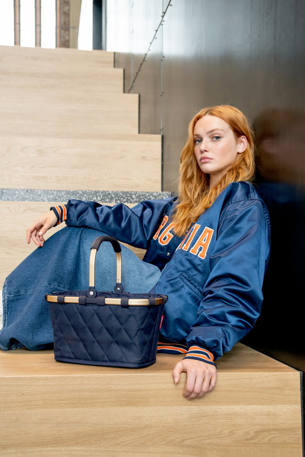 Junge Frau sitzt auf einer Treppe und lehnt sich an die Wand, neben ihr steht eine kleine, gesteppte schwarze Tasche mit hellen Griffen. Perspektive von vorne.