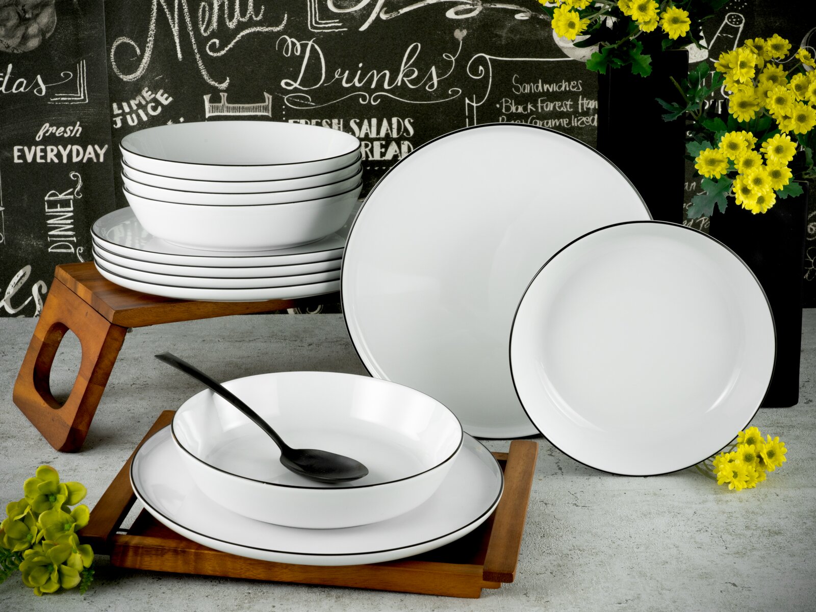 Creatable Tafelservice 12-tlg CHEF COLLECTION Weißes Porzellan-Geschirrset mit schwarzem Rand (Teller und Schalen) auf Holztabletts, fotografiert in schräger Frontansicht vor Tafelhintergrund mit gelben Blumen.