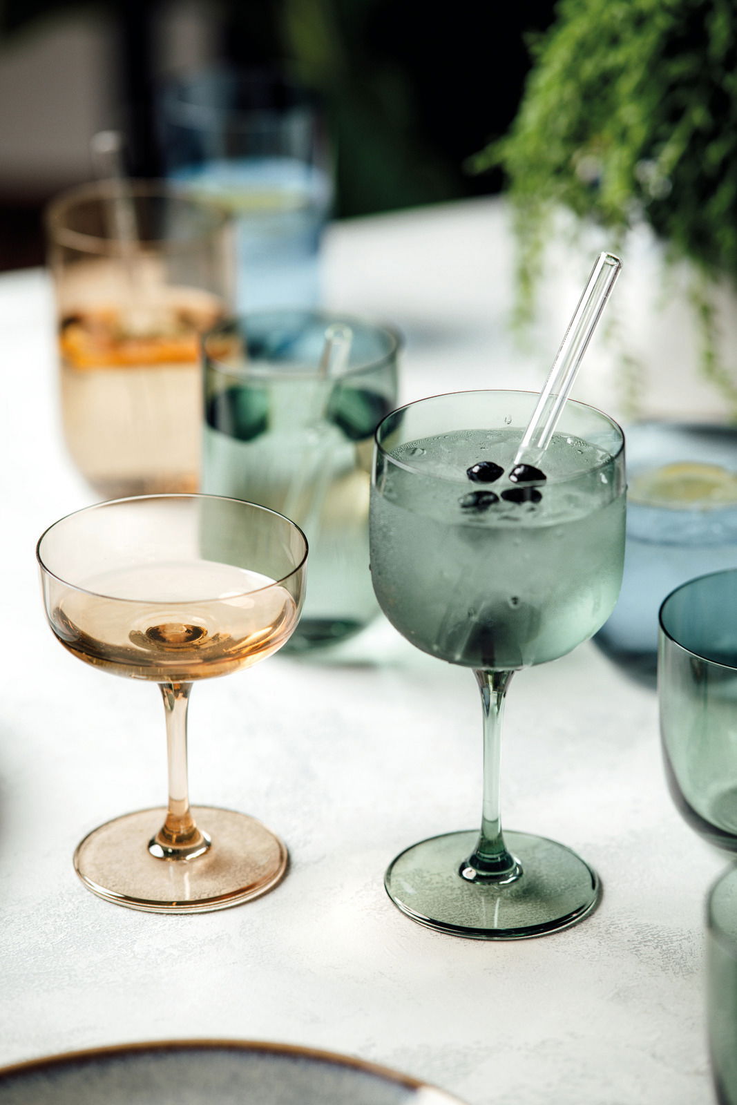 Zwei elegante Weinkelche aus Glas, einer in Bernsteinfarbe und der andere in Grün, auf einem Tisch mit weiteren Gläsern im Hintergrund. Perspektive von schräg oben.