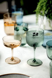 Zwei elegante Weinkelche aus Glas, einer in Bernsteinfarbe und der andere in Grün, auf einem Tisch mit weiteren Gläsern im Hintergrund. Perspektive von schräg oben.