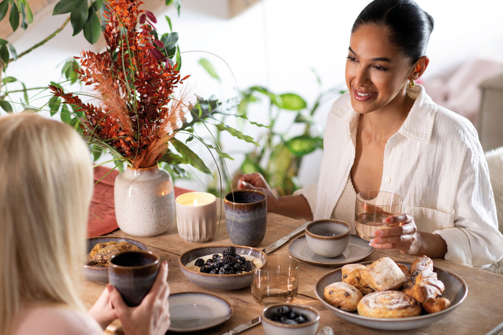 Zwei Personen sitzen an einem Tisch mit einem Wasserglas-Set, umgeben von Geschirr und einer Vase mit roten Blumen. Die Perspektive zeigt eine gemütliche Frühstücksszene von vorne.