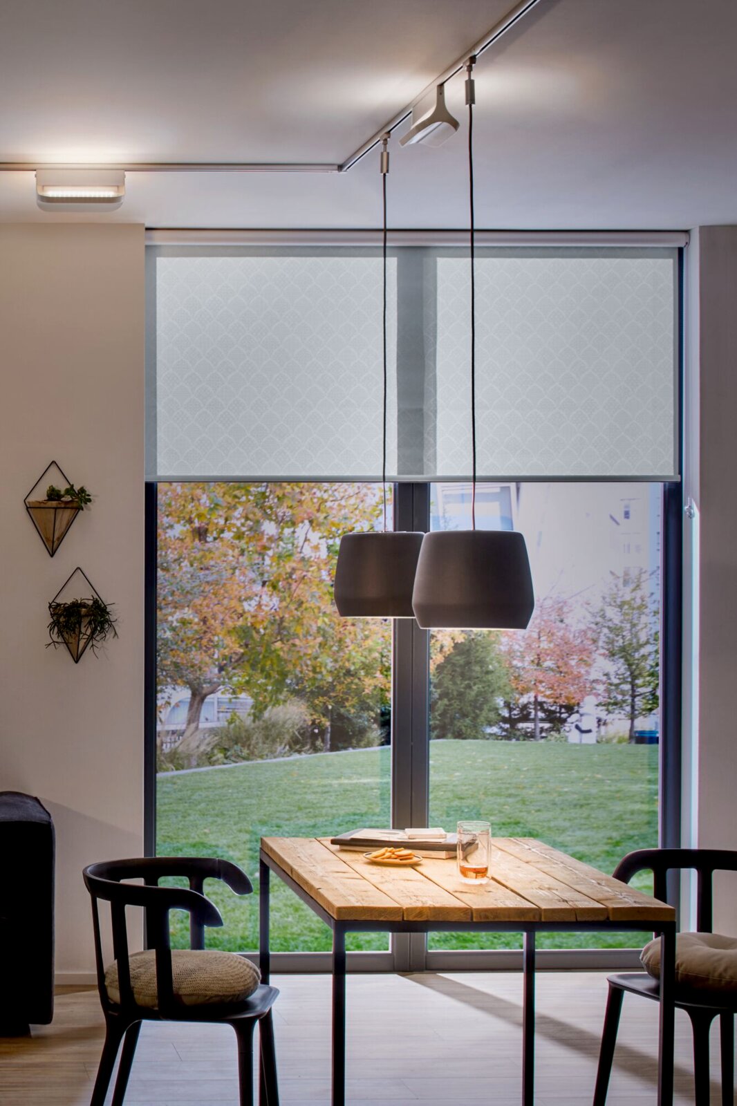 Moderner Innenraum mit hängenden Lampen über einem Holztisch, im Hintergrund ein großes Fenster mit Blick auf einen grünen Garten. Perspektive von vorne.