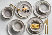 Vogelperspektive auf ein 12-teiliges Tellerset mit grauem, gesprenkeltem Design, bestehend aus Tellern und Schalen, dekoriert mit goldfarbenem Besteck und Lebensmitteln.