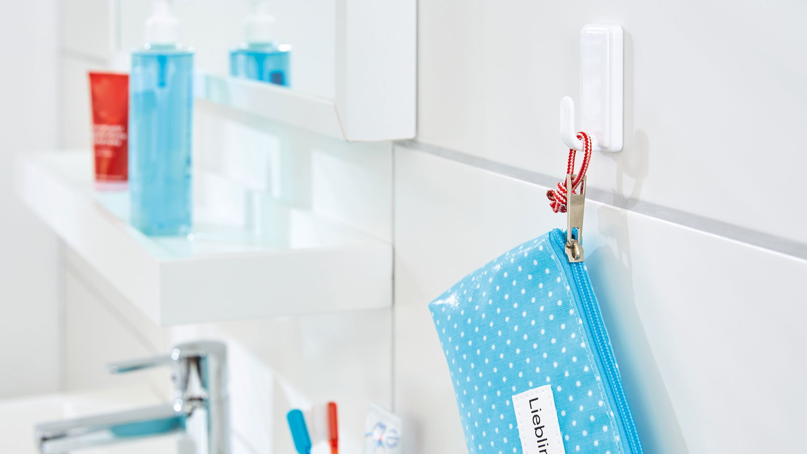Ein weißer tesa Powerstrips Haken Large an einer Badezimmerwand, der eine blaue gepunktete Tasche mit rotem Band hält. Perspektive von der Seite.