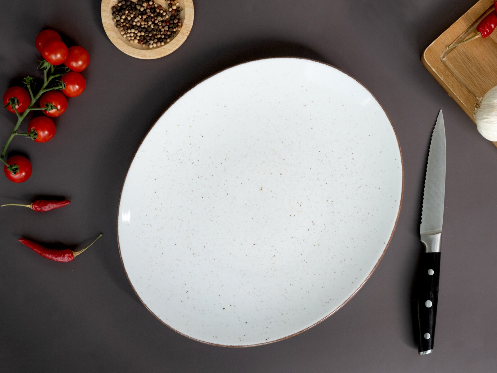 Runder, weiß gesprenkelter Keramikteller mit braunem Rand auf dunkler Oberfläche, umgeben von Kirschtomaten, Chilischoten, Pfefferkörnern, Knoblauch, Schneidebrett und Messer, Draufsicht