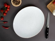 Runder, weiß gesprenkelter Keramikteller mit braunem Rand auf dunkler Oberfläche, umgeben von Kirschtomaten, Chilischoten, Pfefferkörnern, Knoblauch, Schneidebrett und Messer, Draufsicht