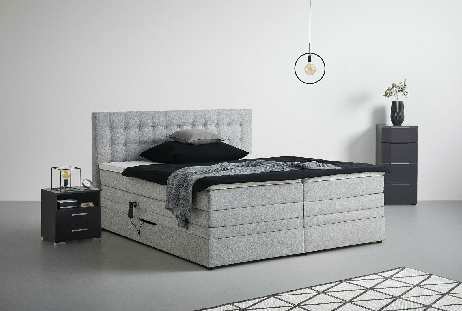 Graues Boxspringbett mit gestepptem Kopfteil, seitlicher Perspektive, umgeben von modernen Nachttischen und minimalistischer Dekoration.
