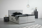 Graues Boxspringbett mit gestepptem Kopfteil, seitlicher Perspektive, umgeben von modernen Nachttischen und minimalistischer Dekoration.