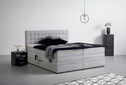 Graues Boxspringbett mit gestepptem Kopfteil, seitlicher Perspektive, umgeben von modernen Nachttischen und minimalistischer Dekoration.