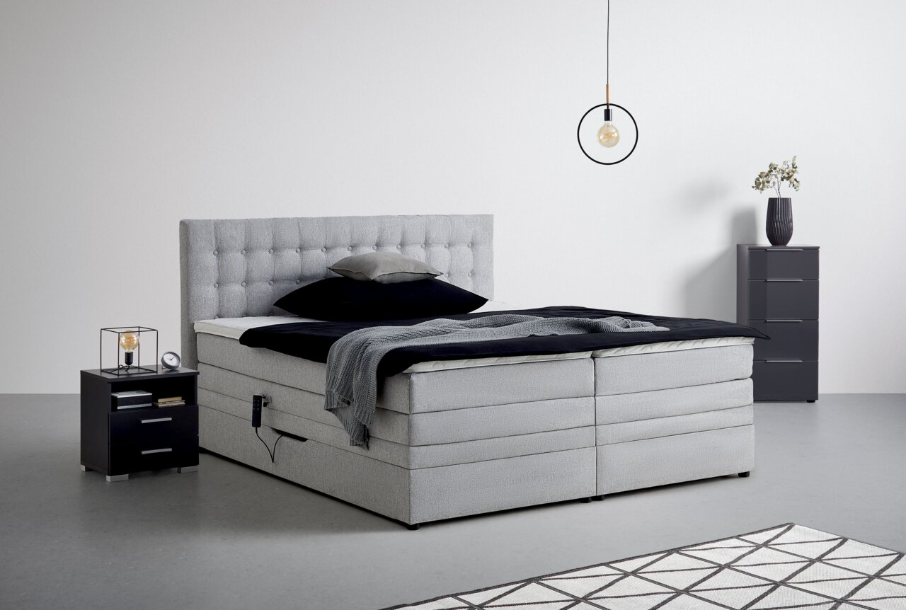 Graues Boxspringbett mit gestepptem Kopfteil, seitlicher Perspektive, umgeben von modernen Nachttischen und minimalistischer Dekoration.