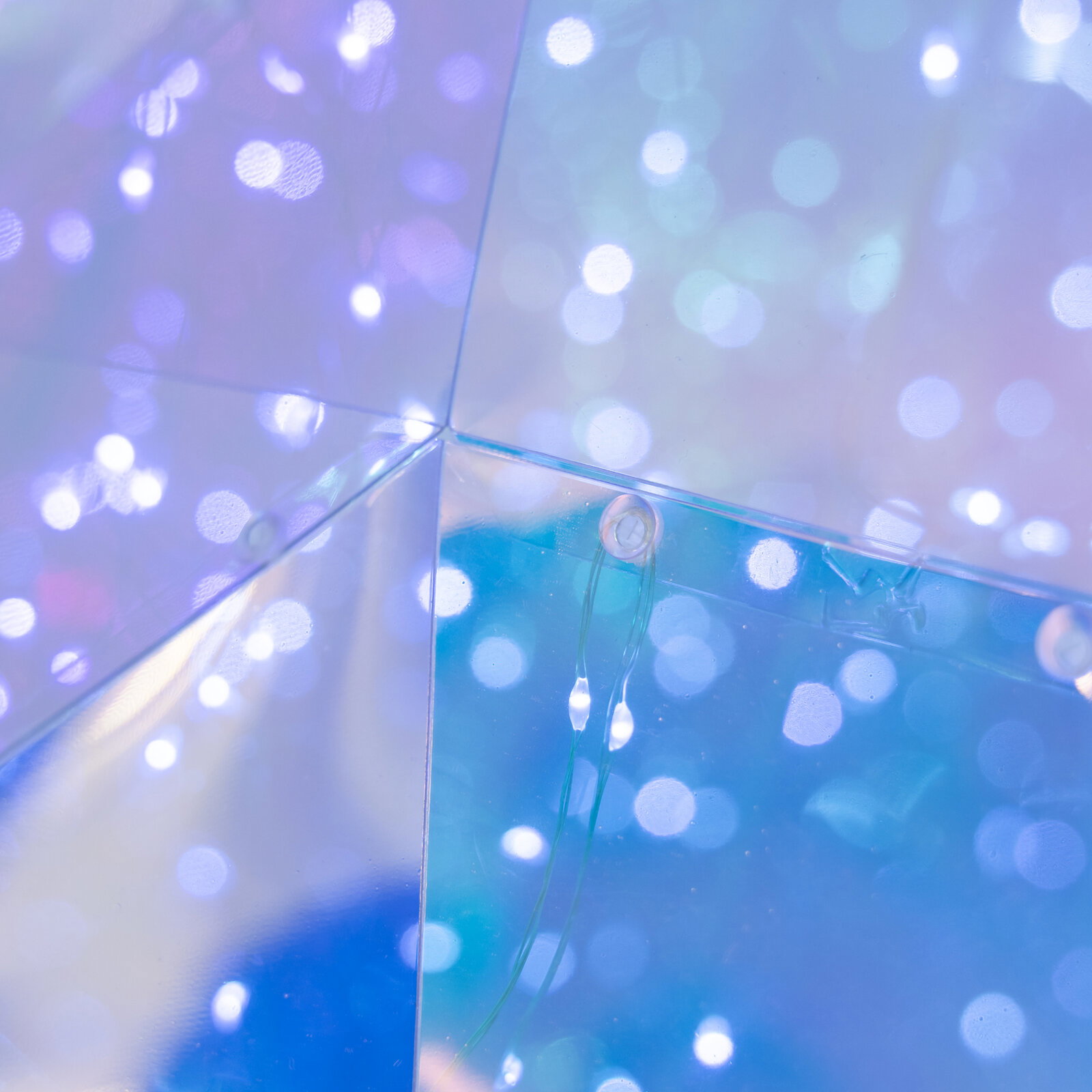 Nahaufnahme aus schräger Innenperspektive auf die Ecke einer transparenten Acrylbox mit blau-violettem Bokeh-Licht und sichtbarer Befestigung/Lasche.