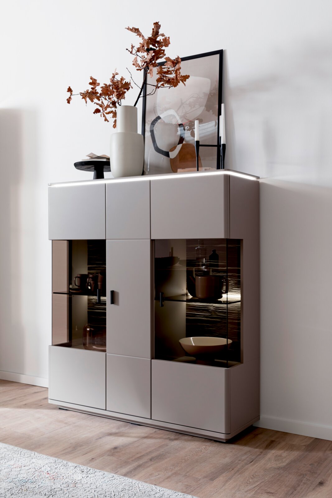 Modernes, hellgraues Sideboard mit zwei beleuchteten Glasvitrinen und schwarzen Griffen, dekoriert mit Vase und Bild, in schräger Frontansicht von links vor weißer Wand.