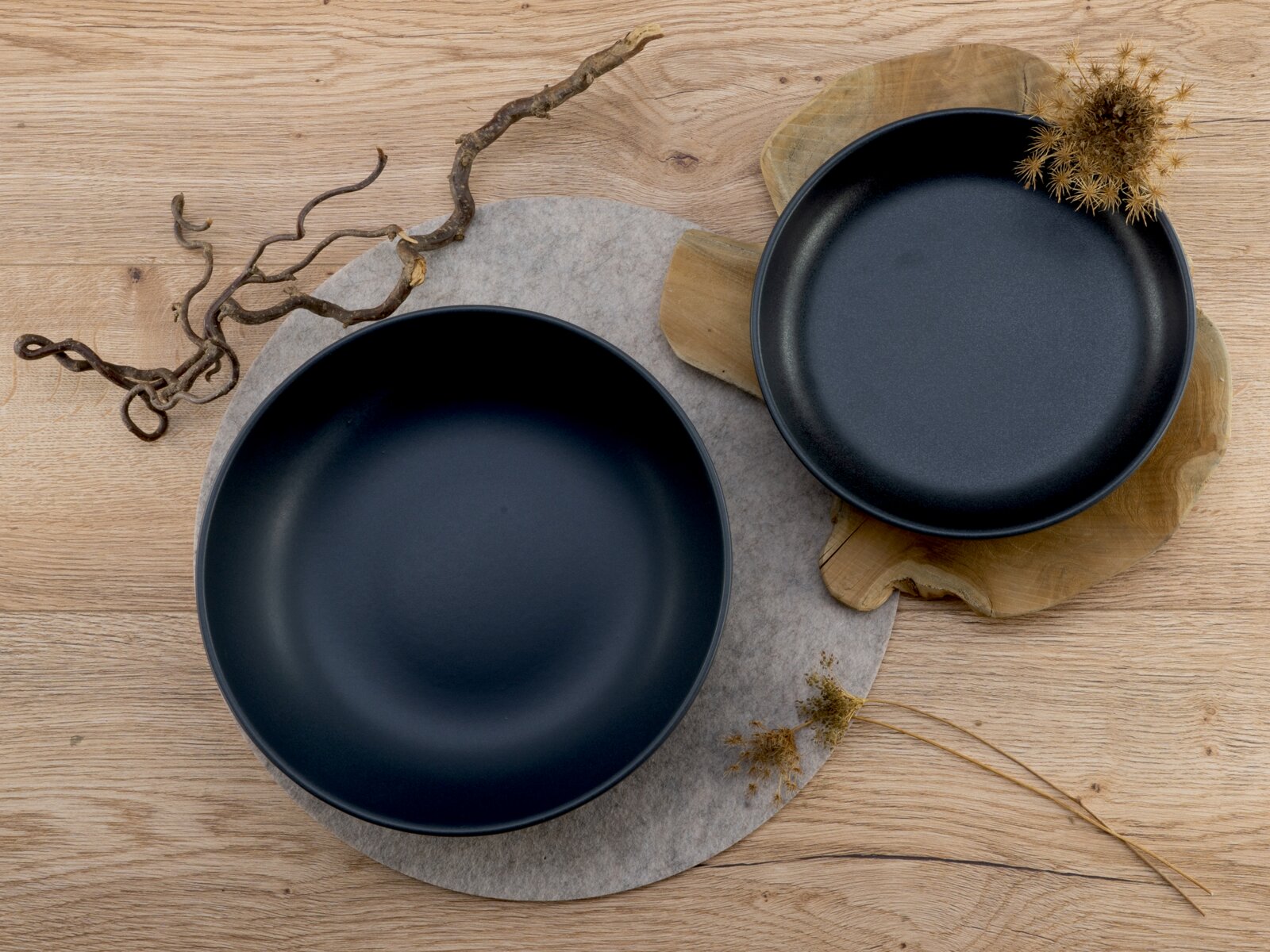 Zwei matte schwarze Keramikschalen auf Holzuntergrund, von oben fotografiert; eine große Schale auf rundem Filzuntersetzer, eine kleinere auf Holzbrett mit Deko-Zweigen.