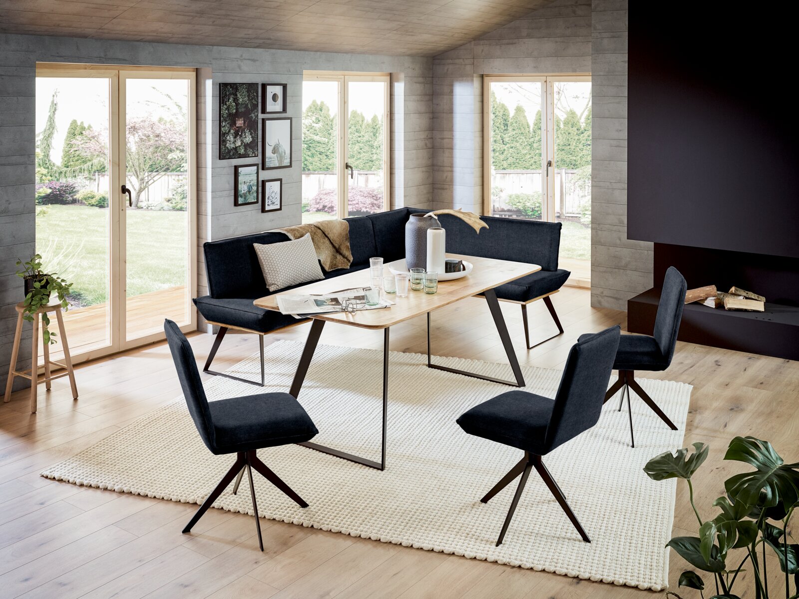 Moderne hoekbankgroep in een lichte woonkamer met grote ramen, bestaande uit een hoekbank en vier stoelen met donkere bekleding, opgesteld rond een houten tafel. Aanzicht van schuin bovenaf.