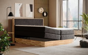 Zijaanzicht van een modern boxspringbed met grijze stoffen bekleding en een houten frame in een stijlvol ingerichte slaapkamer met grote ramen.
